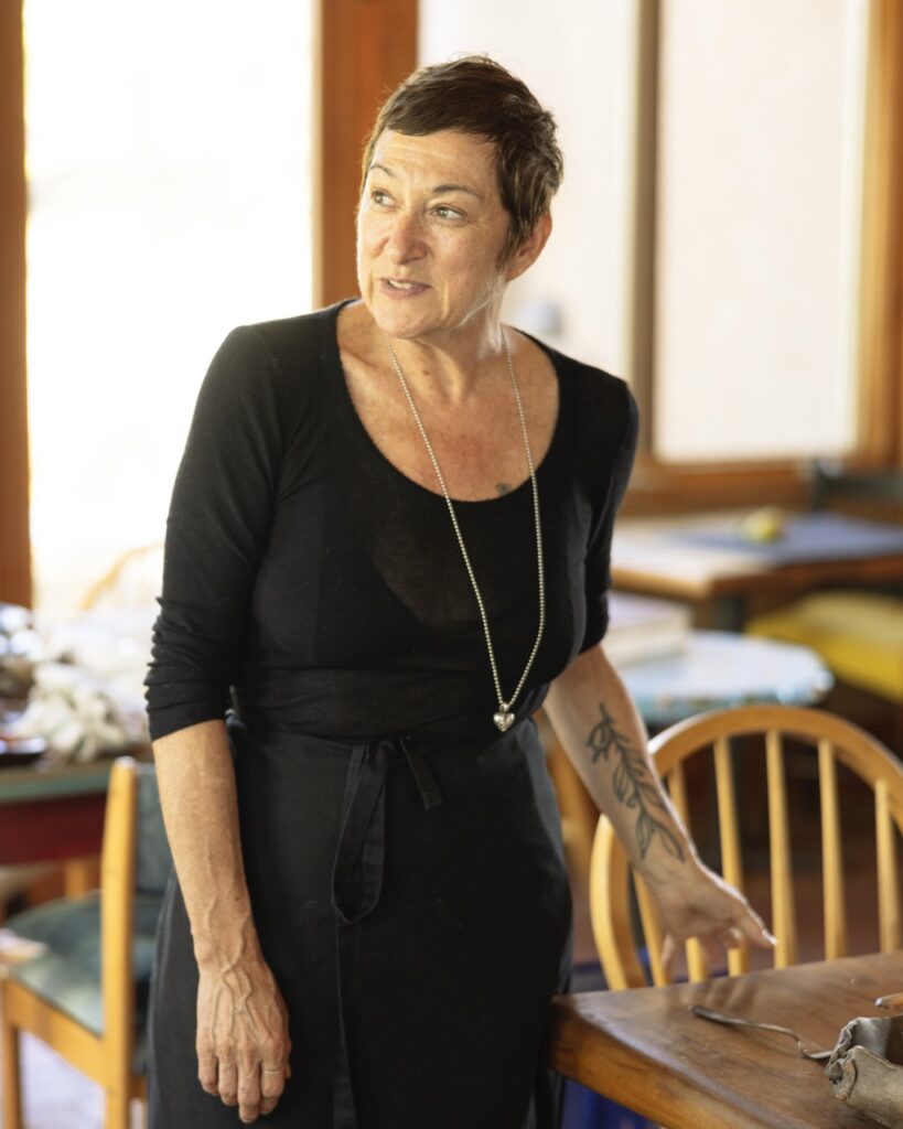 A woman in a black dress with a long silver necklace leans against a wood table.