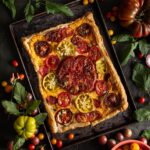 A rectangular heirloom tomato pie with a flaky puff pastry crust, topped with colorful sliced tomatoes and fresh thyme, on a dark baking sheet surrounded by various whole and halved heirloom tomatoes and greenery.