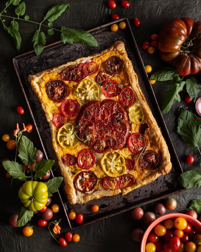 A rectangular heirloom tomato pie with a flaky puff pastry crust, topped with colorful sliced tomatoes and fresh thyme, on a dark baking sheet surrounded by various whole and halved heirloom tomatoes and greenery.