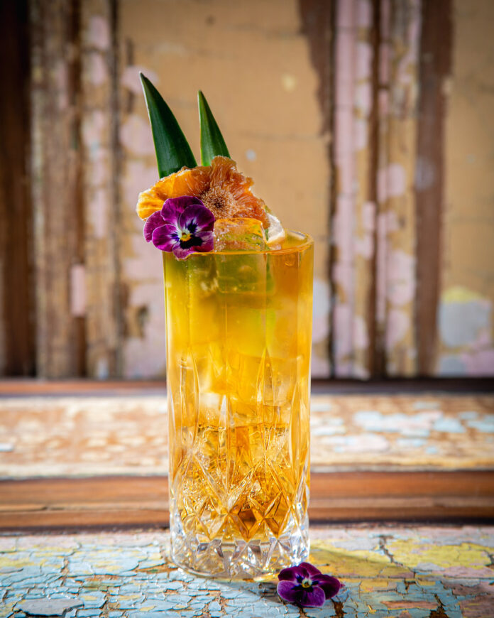 A tropic funder cocktail in a tall, faceted glass is garnished with two green leaves, an orange dehydrated pineapple slice, and a purple edible flower.