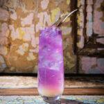 A tall, clear glass holds a layered purple and white unicorn tears cocktail, garnished with several gold-colored, spiky decorative leaves