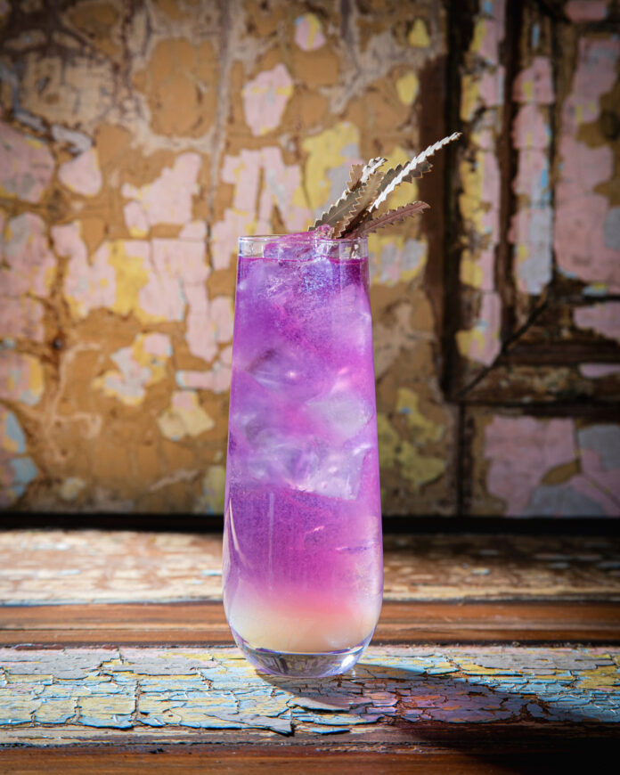 A tall, clear glass holds a layered purple and white unicorn tears cocktail, garnished with several gold-colored, spiky decorative leaves
