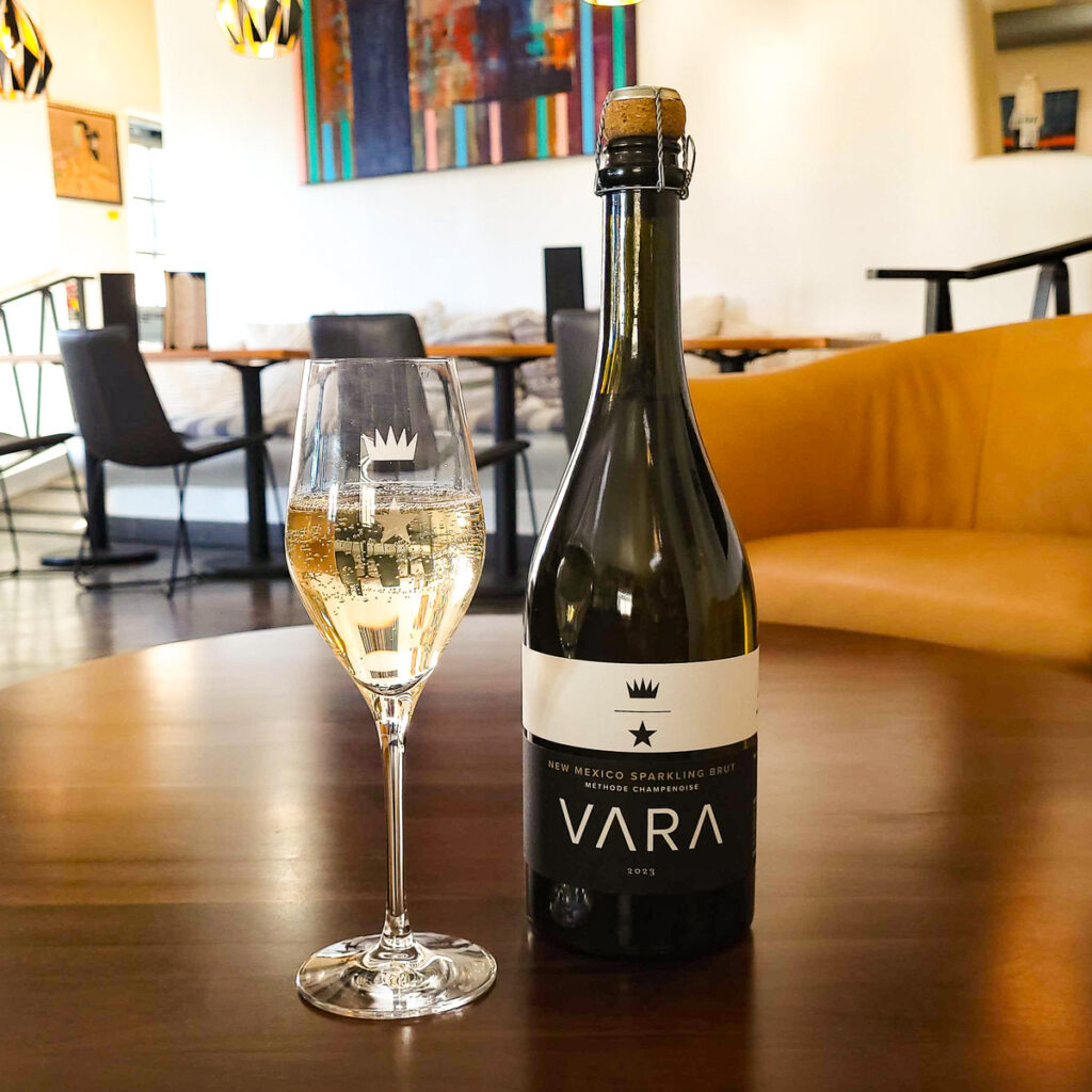 A bottle and a stemmed glass filled with Vara 2023 New Mexico Sparkling Brut wine on a wooden table in a tasting room.
