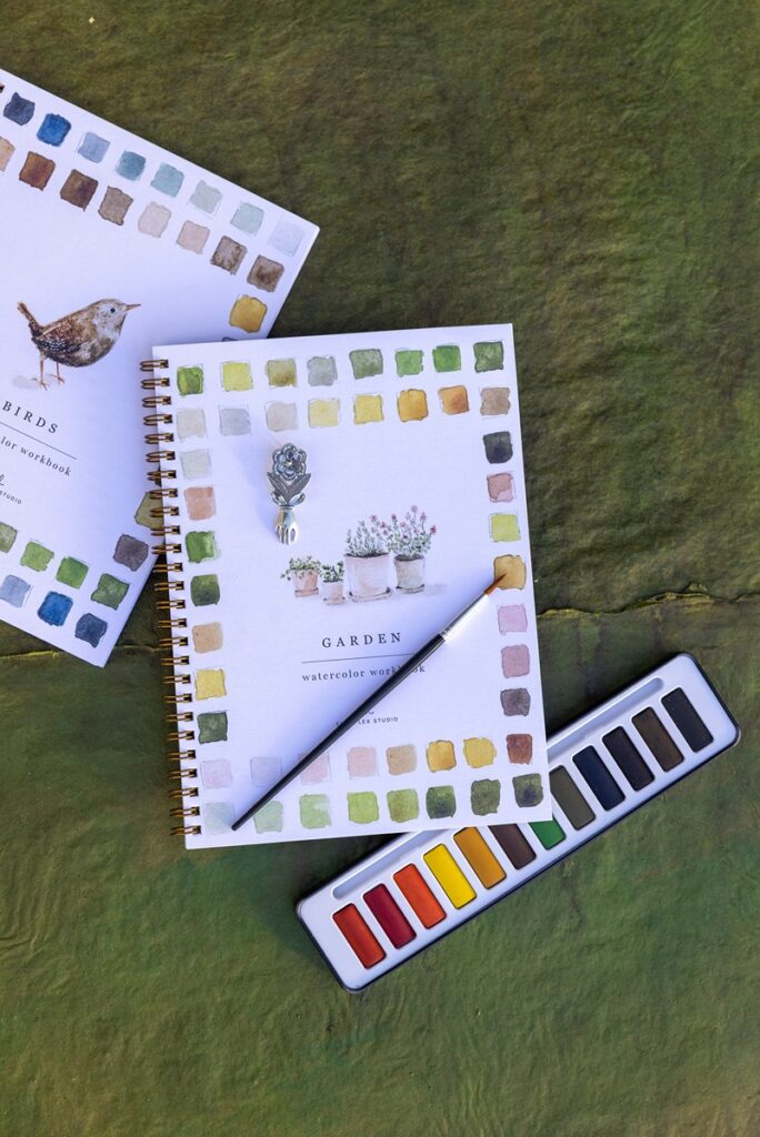 A "Garden" themed watercolor workbook with a spiral binding, a watercolor brush, a paint palette, and a small hand and flower milagro.