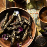 A bowl of Wuzinudeln, Austrian potato noodles coated in poppy seeds and powdered sugar, is garnished with violas and sitting on a rustic wooden surface.