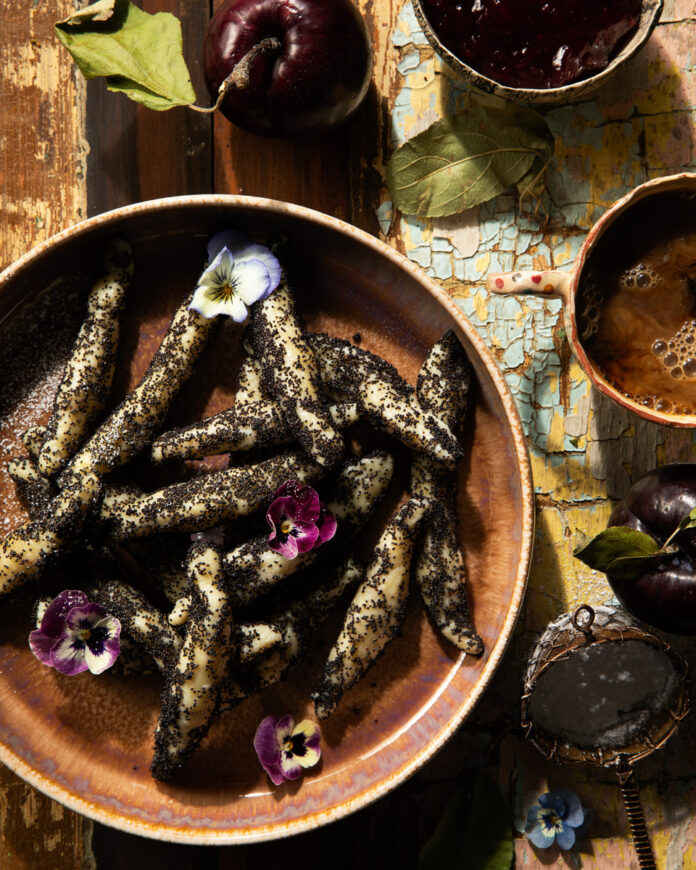 A bowl of Wuzinudeln, Austrian potato noodles coated in poppy seeds and powdered sugar, is garnished with violas and sitting on a rustic wooden surface.