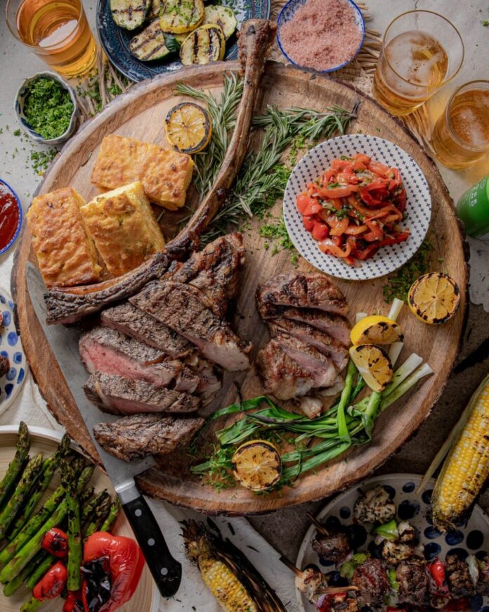 A brown platter holds various summer grilling items such as cornbread, steak, asparagus, corn, peppers, and much more fresh ingredients spread around the platter.