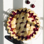 A whole Cherry Pie sits on a white table with a serving knife below the dish and two cherries in the right corner.