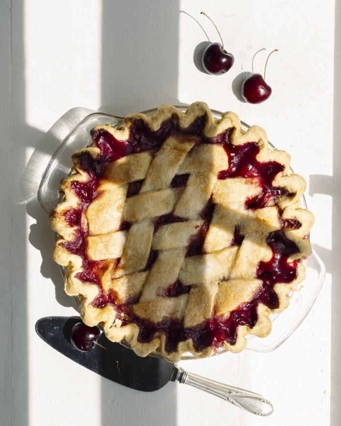 A whole Cherry Pie sits on a white table with a serving knife below the dish and two cherries in the right corner.