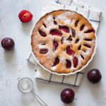 A plum kuchen with plums peaking through the dough coverings and sitting on top of a kitchen towel with plums throughout the table.