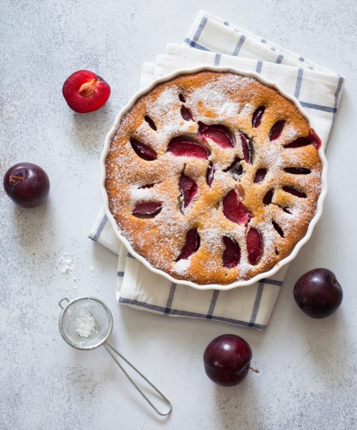 A plum kuchen with plums peaking through the dough coverings and sitting on top of a kitchen towel with plums throughout the table.