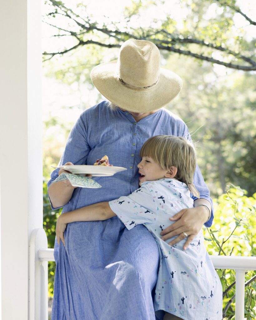 A little boy hugs his mother wearing a long blue dress and sunhat.