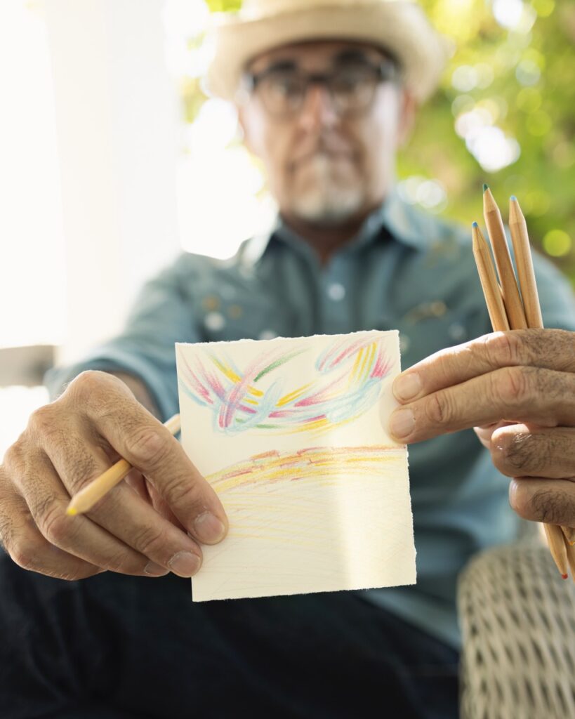 A man in a blue button up holds up a small drawing on a piece of paper with colored pencils.