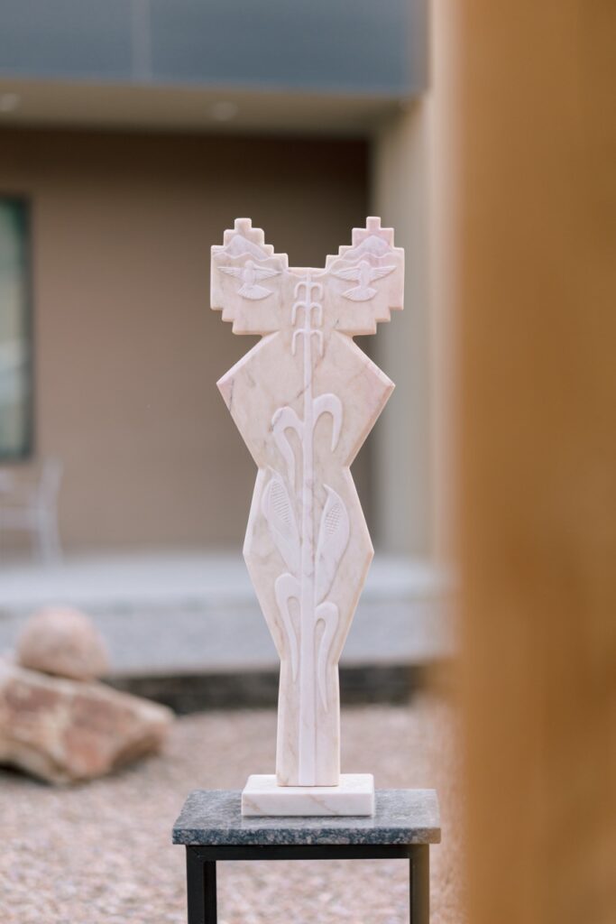 A tall, slender, light-colored abstract sculpture featuring corn and bird motifs.