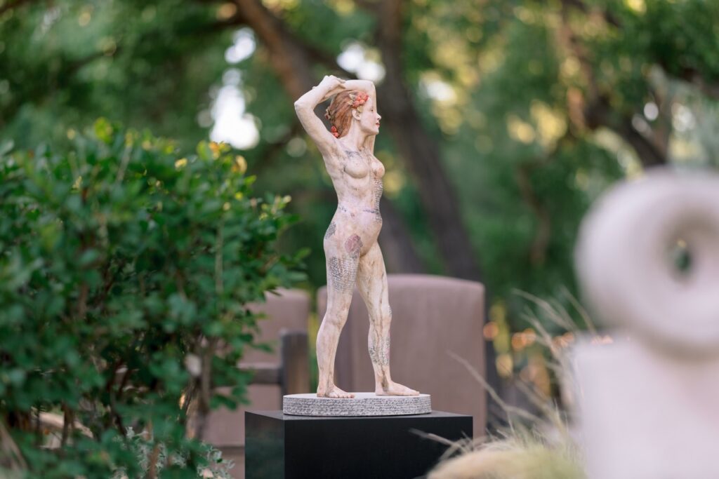 A full-body sculpture of a nude female figure with arms raised, adorned with textured patterns.