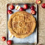 A rustic apple pie, featuring a crust adorned with a Zia symbol and decorative strips, rests on a baking sheet surrounded by whole and halved red apples, all set on a patterned cloth.