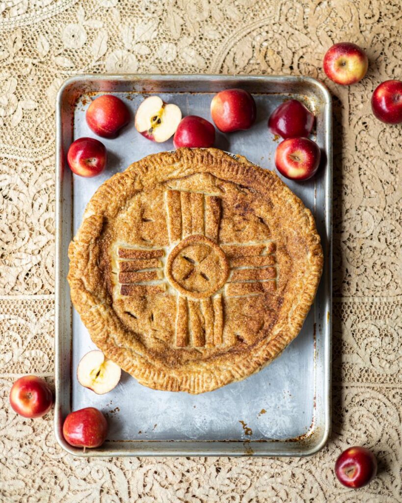 A rustic apple pie, featuring a crust adorned with a Zia symbol and decorative strips, rests on a baking sheet surrounded by whole and halved red apples, all set on a patterned cloth.