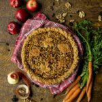 Autumn harvest pie, featuring a golden-brown crust with leaf-shaped cutouts, rests on a red and white checkered cloth, surrounded by fresh apples, carrots, walnuts, and raisins.
