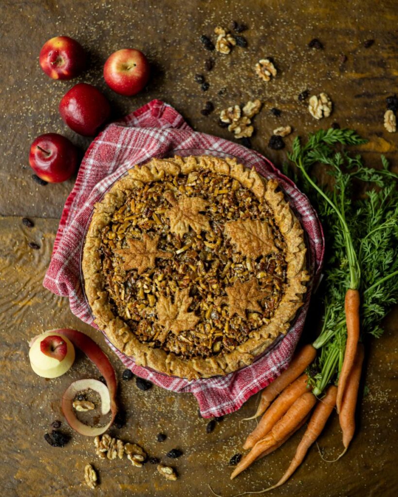 Autumn harvest pie, featuring a golden-brown crust with leaf-shaped cutouts, rests on a red and white checkered cloth, surrounded by fresh apples, carrots, walnuts, and raisins.
