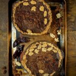 Two freshly baked pecan pies, adorned with decorative pastry leaves, are on a baking sheet, with dried red chiles and scattered pecan halves beside them.