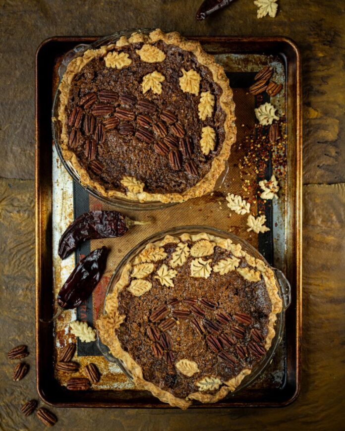Two freshly baked pecan pies, adorned with decorative pastry leaves, are on a baking sheet, with dried red chiles and scattered pecan halves beside them.