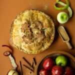 A freshly baked apple pie with a golden-brown crust and decorative leaf cutouts rests on an orange surface, surrounded by red and green apples, cinnamon sticks, apple peels, and a scoop of brown sugar.