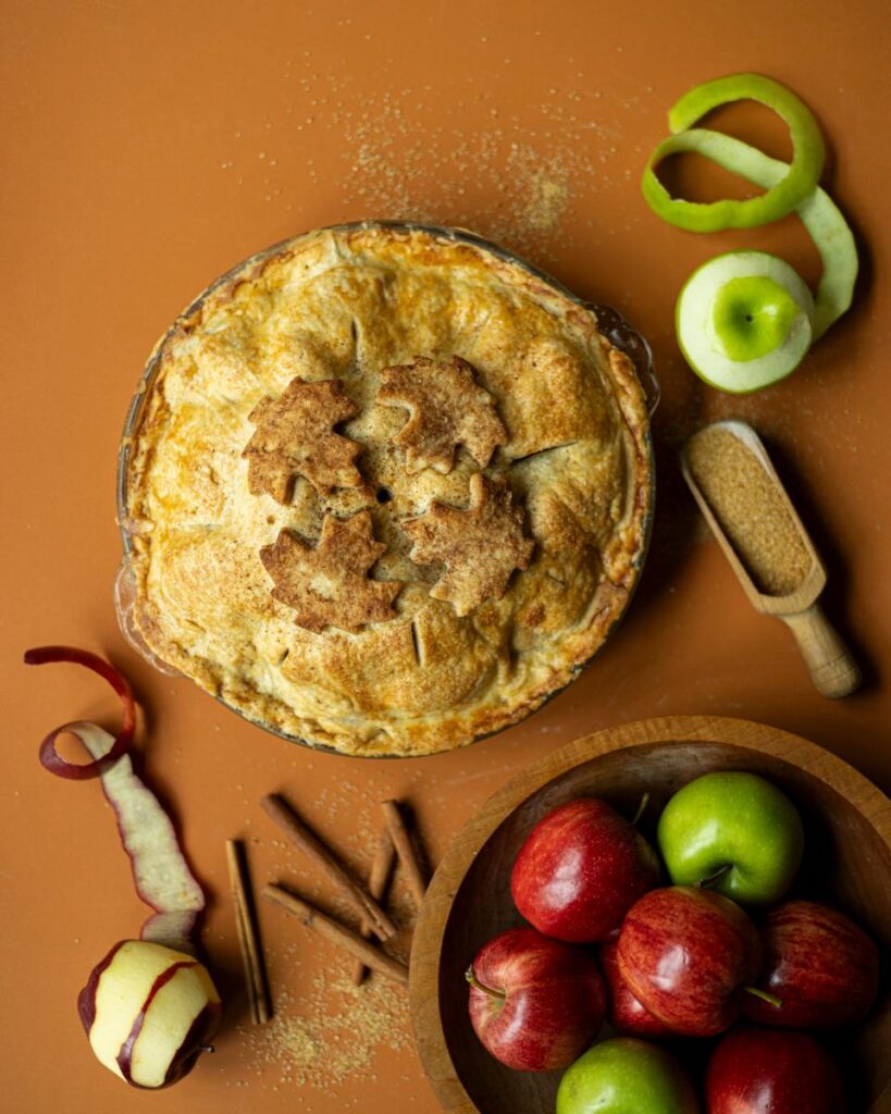 A freshly baked apple pie with a golden-brown crust and decorative leaf cutouts rests on an orange surface, surrounded by red and green apples, cinnamon sticks, apple peels, and a scoop of brown sugar.