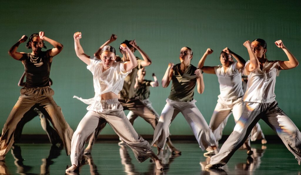 A group of dancers from the Zeitgeist Dance Theatre dance on a green stage in white outfits.