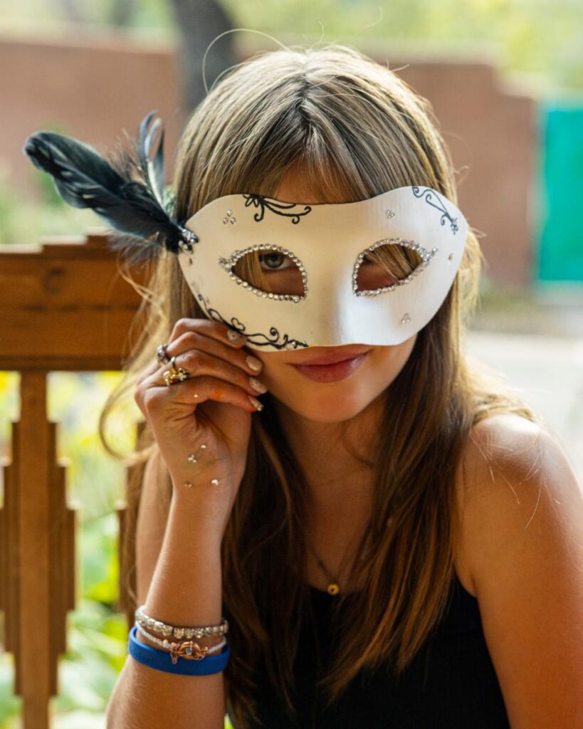 A girl with a white and black mask over her eyes is holding a small diamond-like sticker in her hand to add to the mask