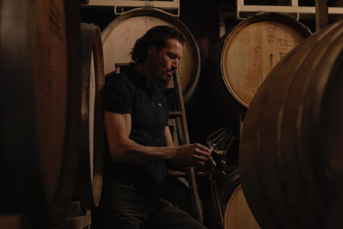 A man with long, dark hair is inspecting a glass of wine, with several large wooden wine barrels visible in the background.