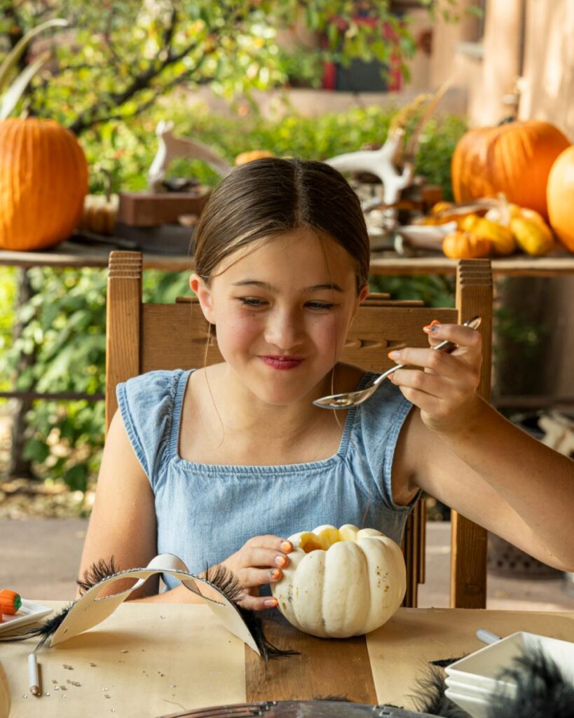 A teenage girl in a denim dress is sitting at a wooden table, smiling while eating soup out of a hollowed-out pumpkin with a silver spoon.