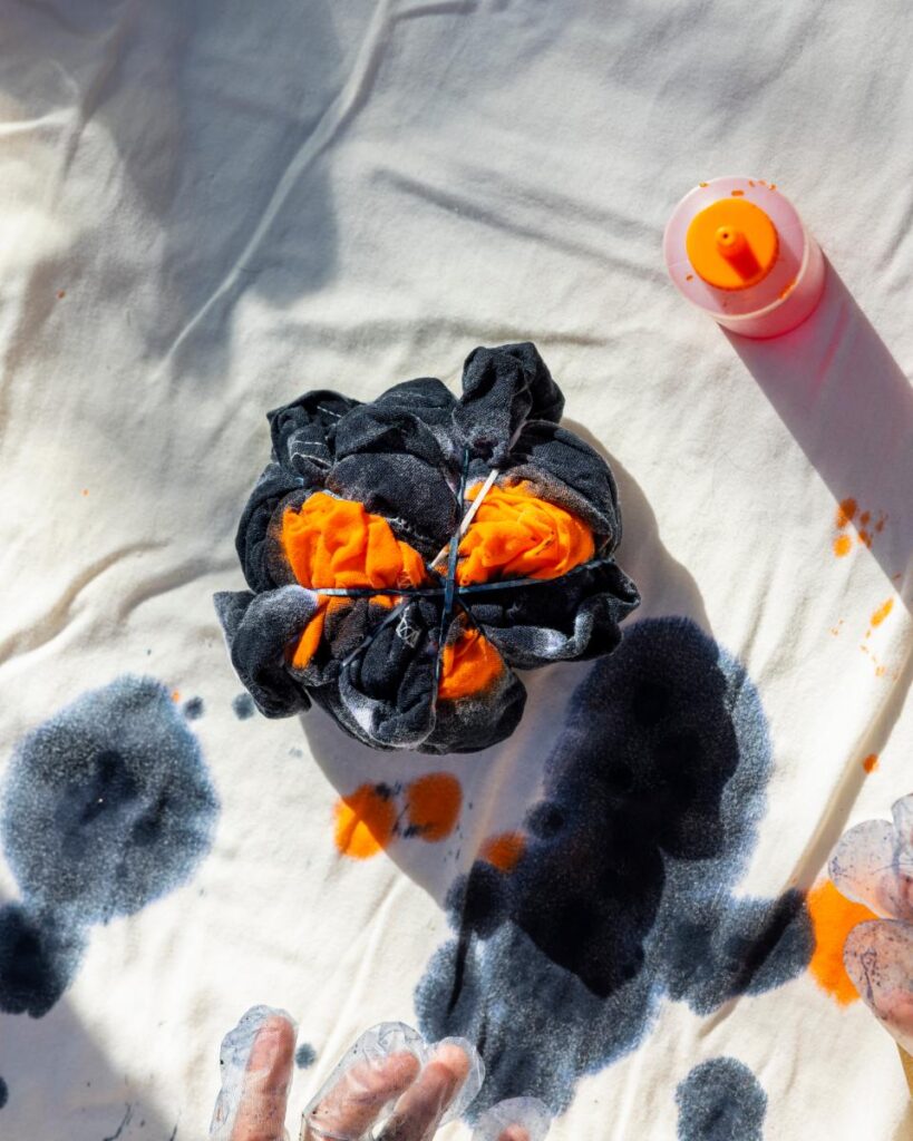 A black cloth is tied with rubber bands and soaked in orange and black dye on a white cloth surface.