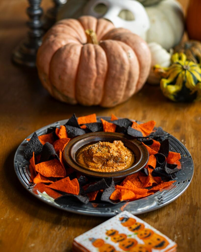 A plate of red pepper hummus topped with seeds is surrounded by orange and black tortilla chips, with a large, textured pumpkin and gourds in the background.