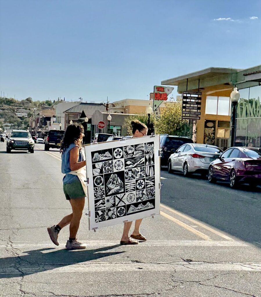 Two women are carrying a large, black and white linoleum print across a street in a downtown area.