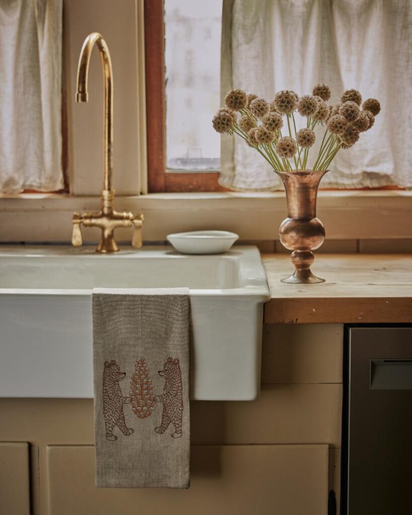 A kitchen sink is shown with a gold faucet and a towel embroidered with two bears holding a tree, with a vase of dried flowers on the counter.
