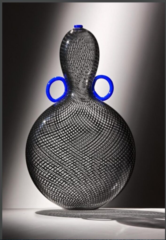 A large glass vase with a black and white net pattern on a white surface, with two bright blue rings on the top sides of the vase.