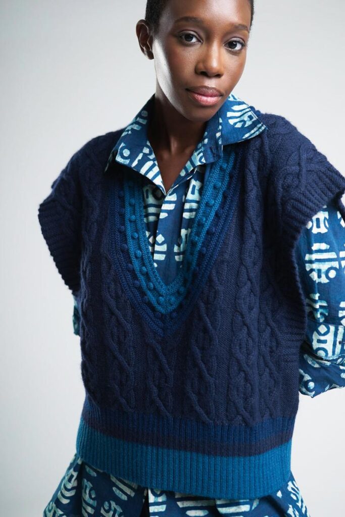 A black woman wearing a navy blue cable knit sweater vest over a light blue patterned long-sleeve blouse is looking at the camera.