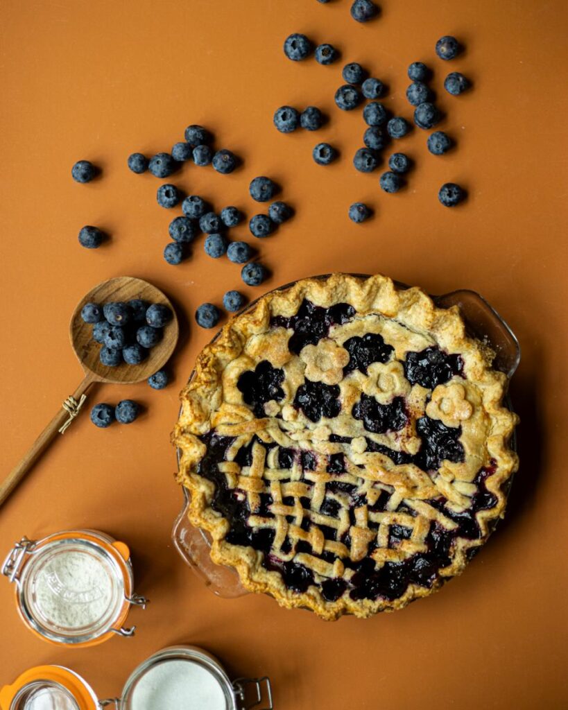 A blueberry pie with a decorative latticed crust is on a wooden surface, with a wooden spoon holding fresh blueberries.