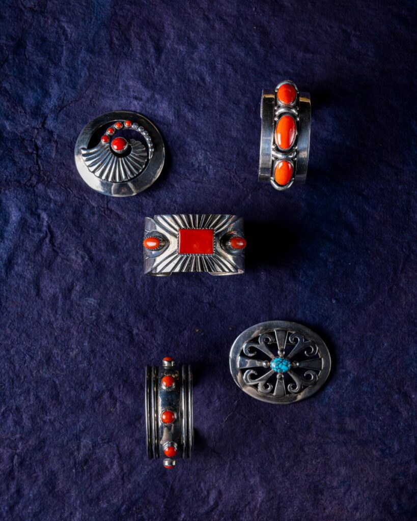 A top-down view of five vintage silver and coral jewelry pieces, including two bracelets and three pins, arranged on a dark blue background.
