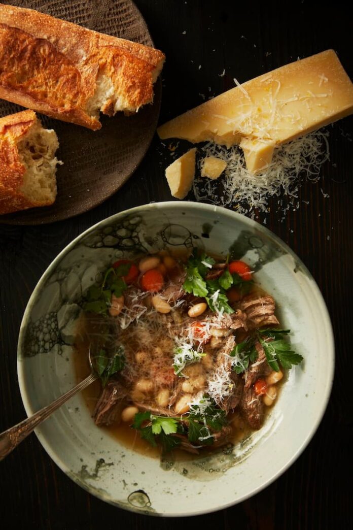 A bowl of brodo di carne soup with shredded beef, cannellini beans, and carrots, topped with fresh parsley and grated parmesan cheese, with bread and a wedge of parmesan on the side.