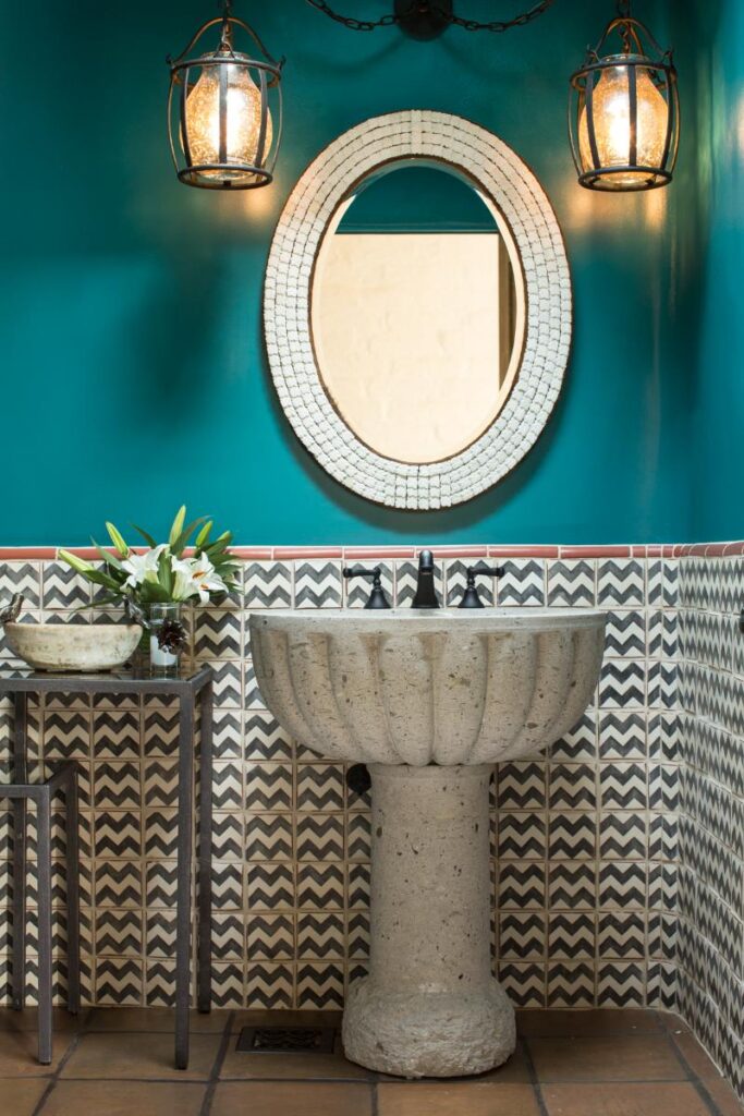 A powder room with vibrant turquoise-colored walls. A white oval mirror is mounted on the wall with black lamps on either side. A carved, gray, stone pedestal sink sits below the mirror.