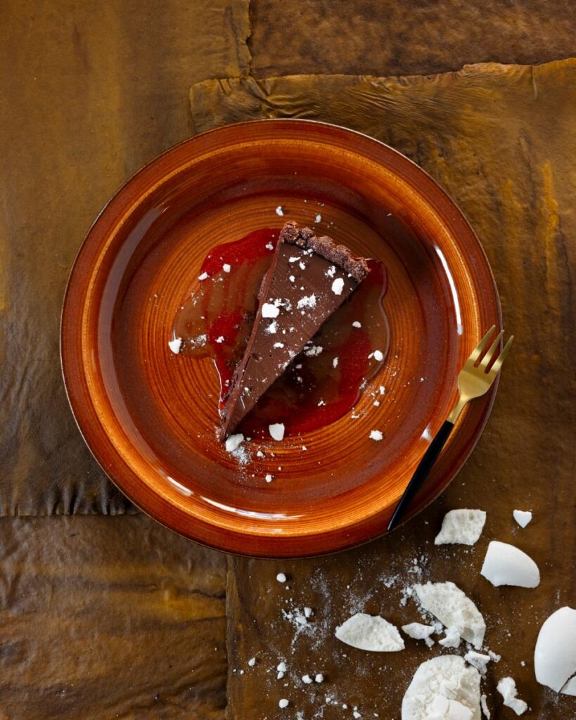 A slice of rich chocolate torte is topped with powdered sugar and drizzled with a red sauce and almond caramel on a reddish-brown plate.