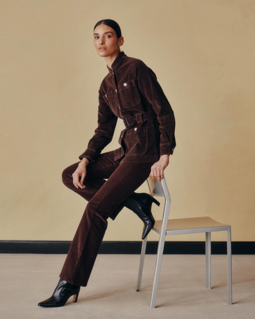 A brown corduroy utility jacket and slim boot-cut pants are shown on a woman sitting on a metal chair.