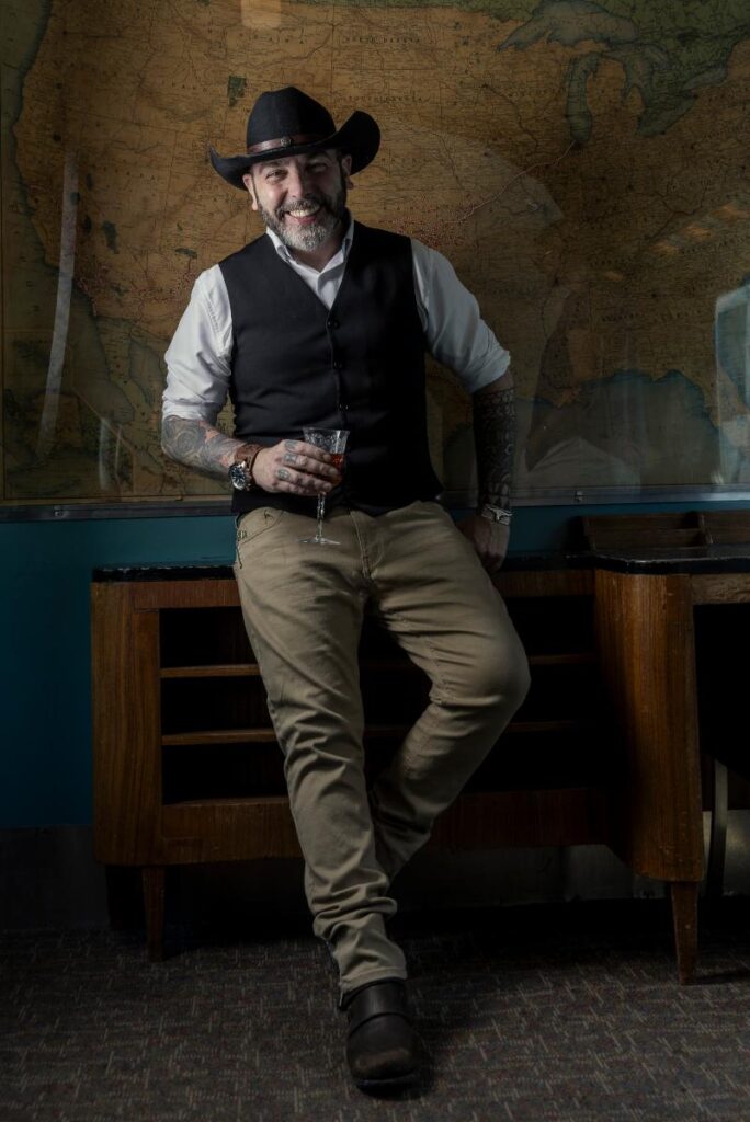 Cristian Pontiggia wearing a cowboy hat and a vest is leaning against a wooden counter, holding a cocktail glass.