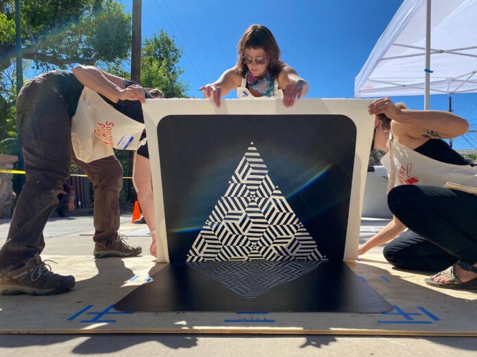 A group of people are gathered around a large piece of art on the ground, with two people on either side of the print lifting it to reveal a black and white geometric pattern.