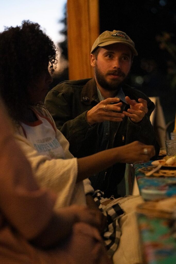 A man with a beard and a baseball cap, sitting at a dinner table and talking, with his hands gesturing.
