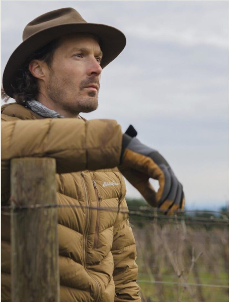 Evan Martin is leaning against a barbed wire fencepost, looking off into the distance.