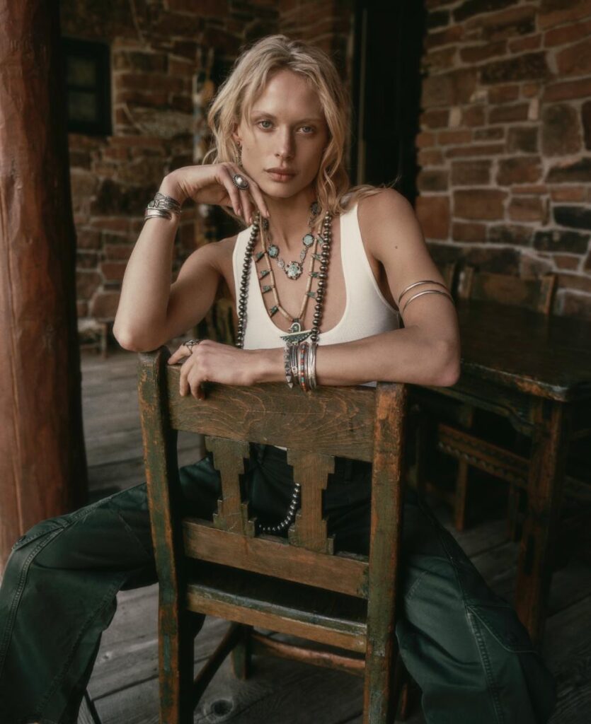 A fashion portrait of a blonde woman with silver jewelry and a white tank top, sitting backward on a wooden chair with her elbow resting on the back of the chair.