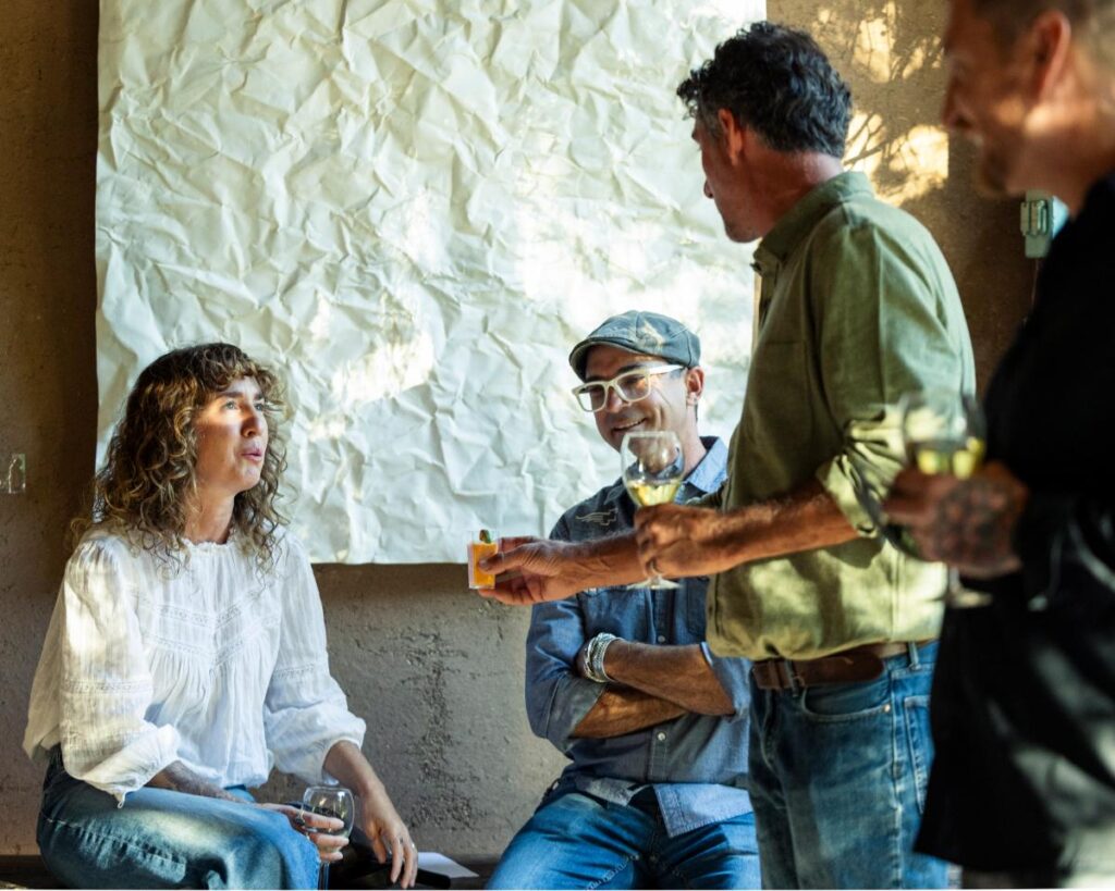 A medium shot of four people standing around, talking, and laughing in a sun-drenched room.