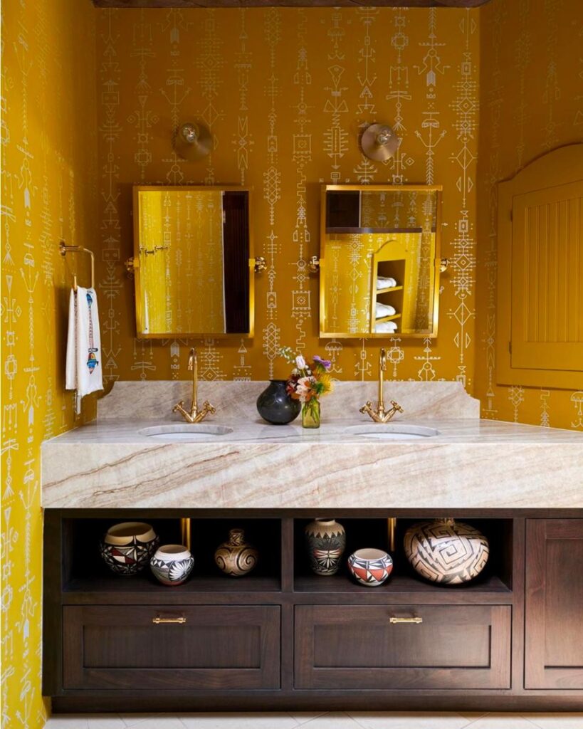 A bathroom with bright yellow patterned wallpaper. A marble countertop with two sinks and brass faucets sits atop a dark wooden vanity with multiple open shelves holding decorated ceramic pots.